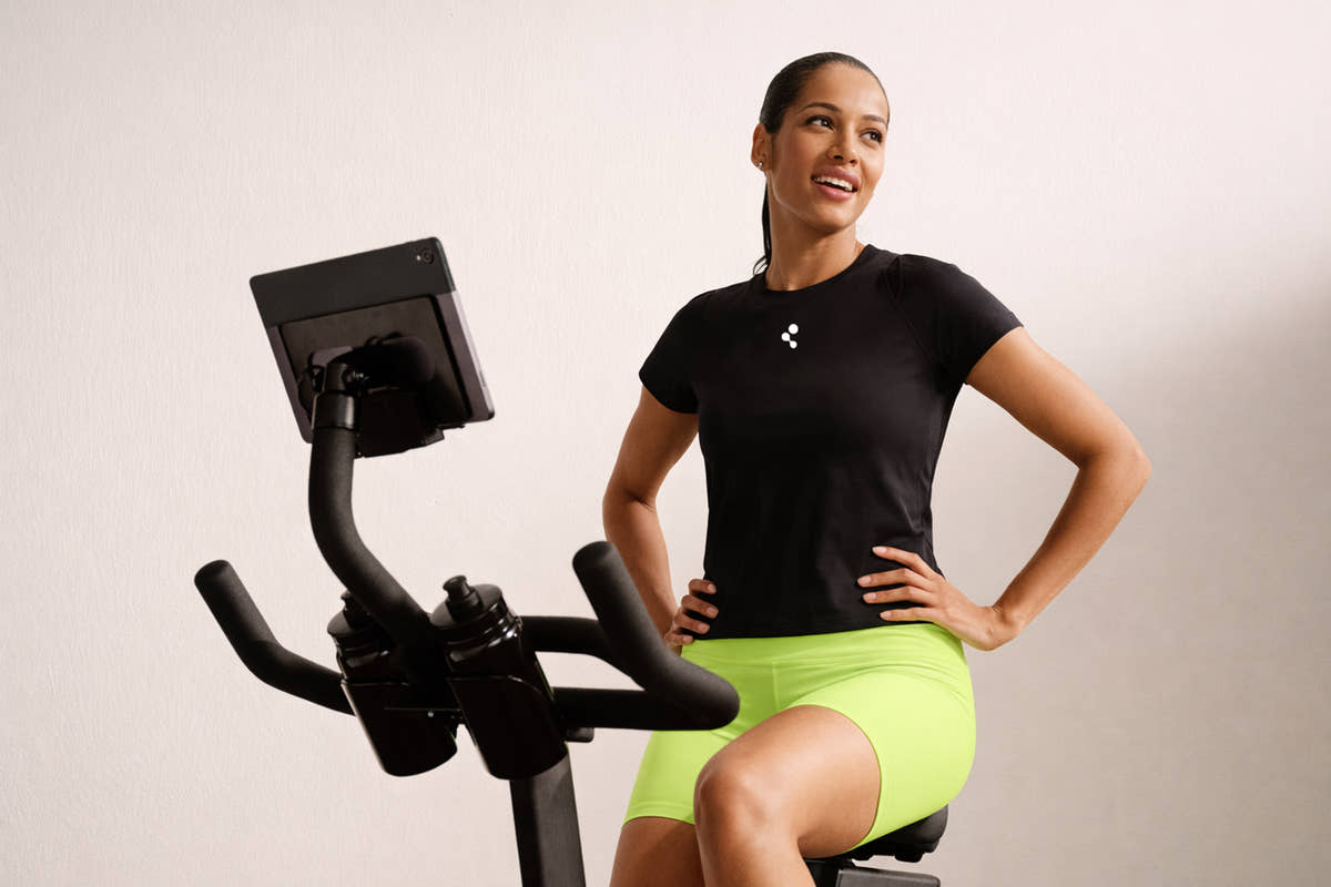 A woman in a black t-shirt sitting beside an exercise bike in a bright studio, hands on her hips
