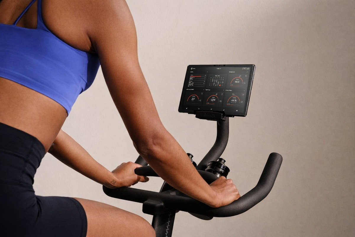 A rider in a blue sports top with her hands on the handlebars of a CAROL bike, the dashboard showing her ride stats